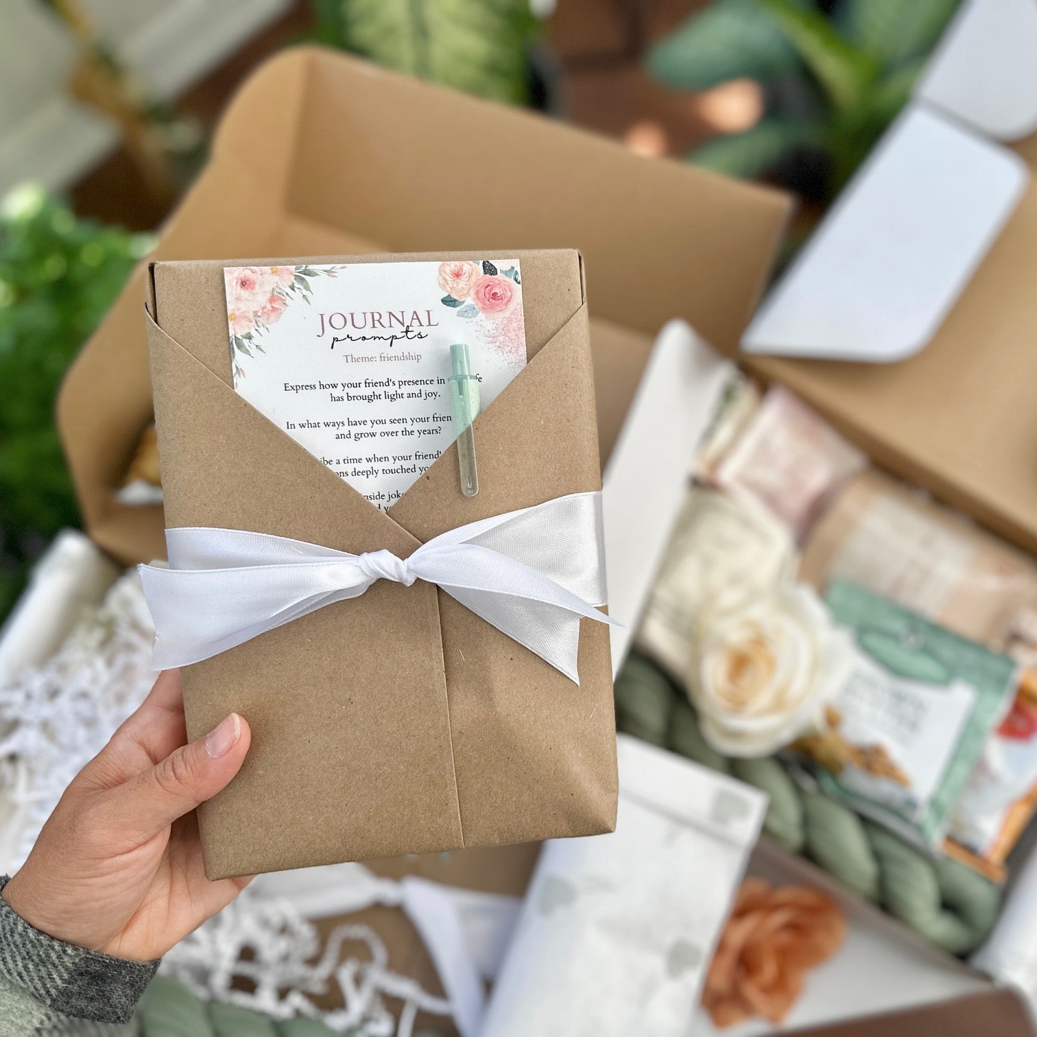 hand holding a bundle of journal that is wrapped in kraft paper, with a journaling prompts and eco pen bundled with it. The background shows other Pen To Paper Experience DIY Kits