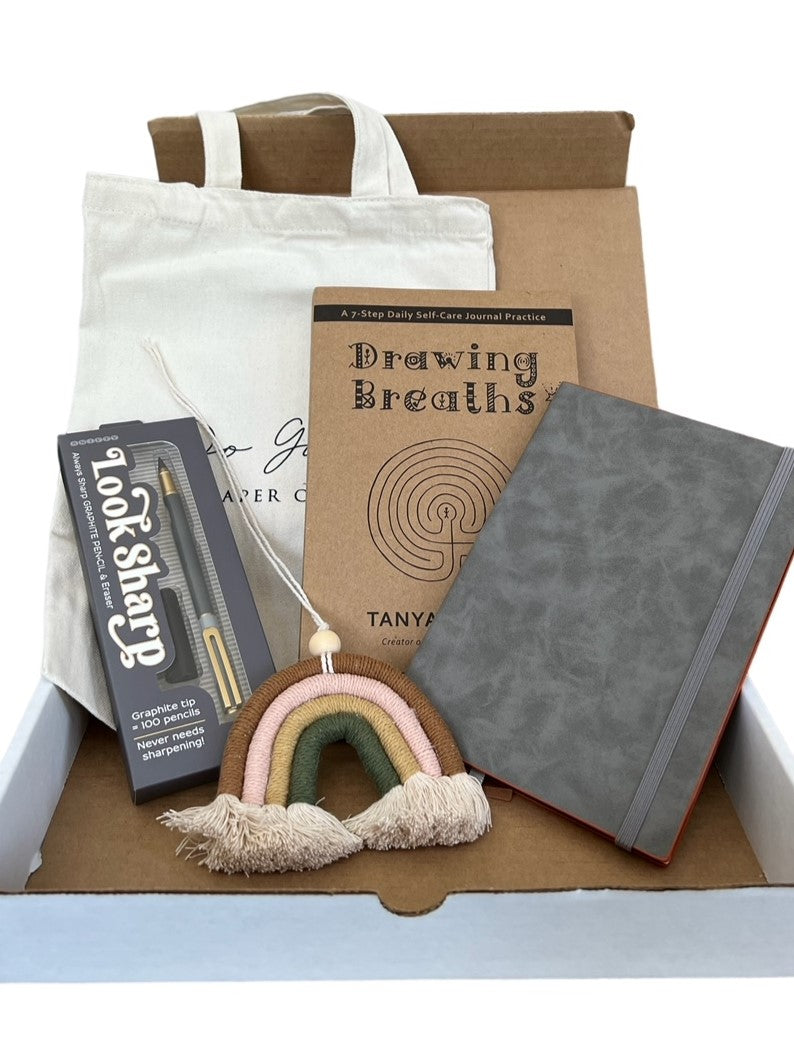 Open stationery subscription box displaying a canvas tote bag, a Drawing Breaths book, a Look Sharp graphite pencil, a grey faux leather journal and a macrame rainbow