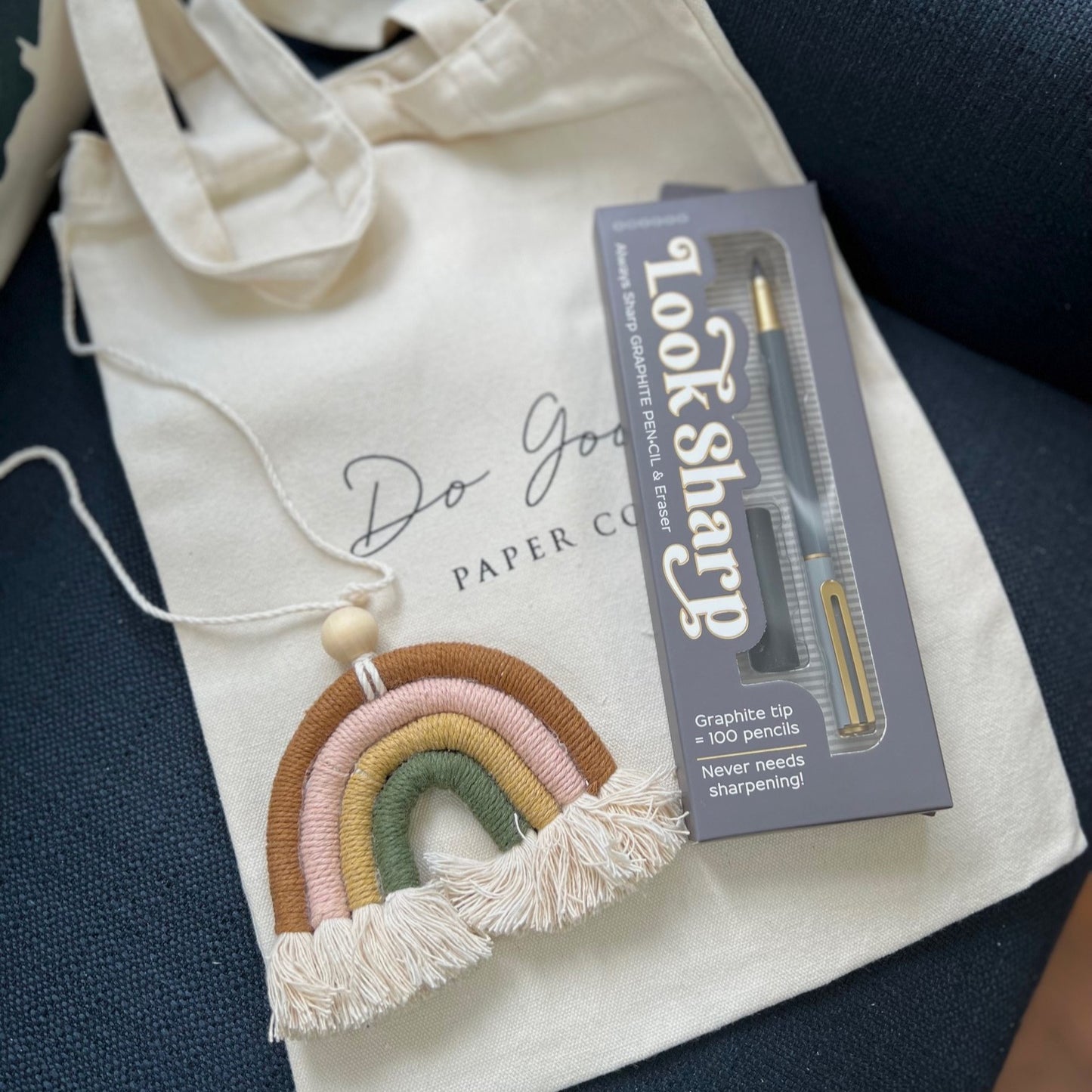 A macrame rainbow and graphite pencil on top of a canvas tote bag displayed on a dark blue fabric chair