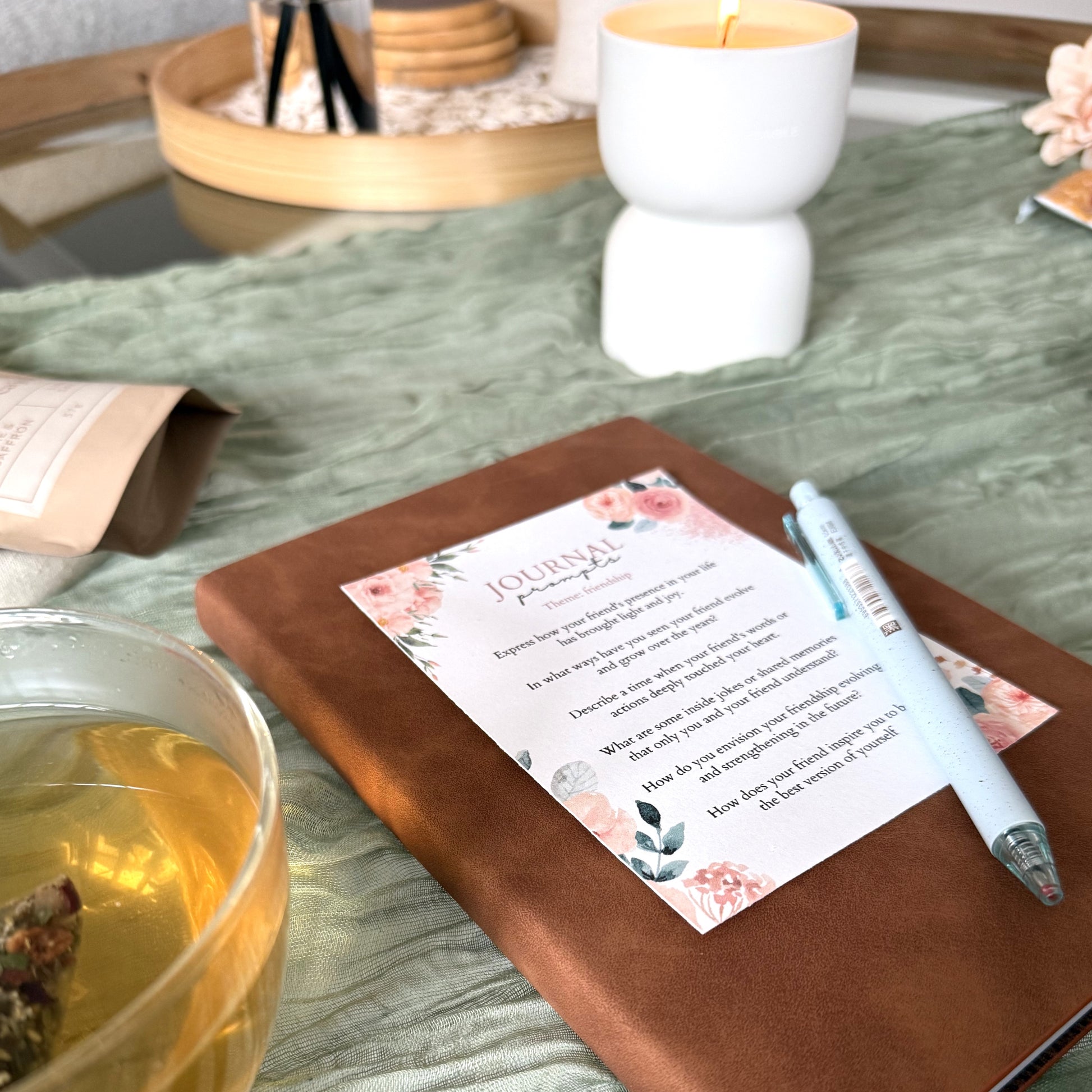 brown vegan leather journal with writing prompts card and eco pen on top of it, lying on a table with green table cloth