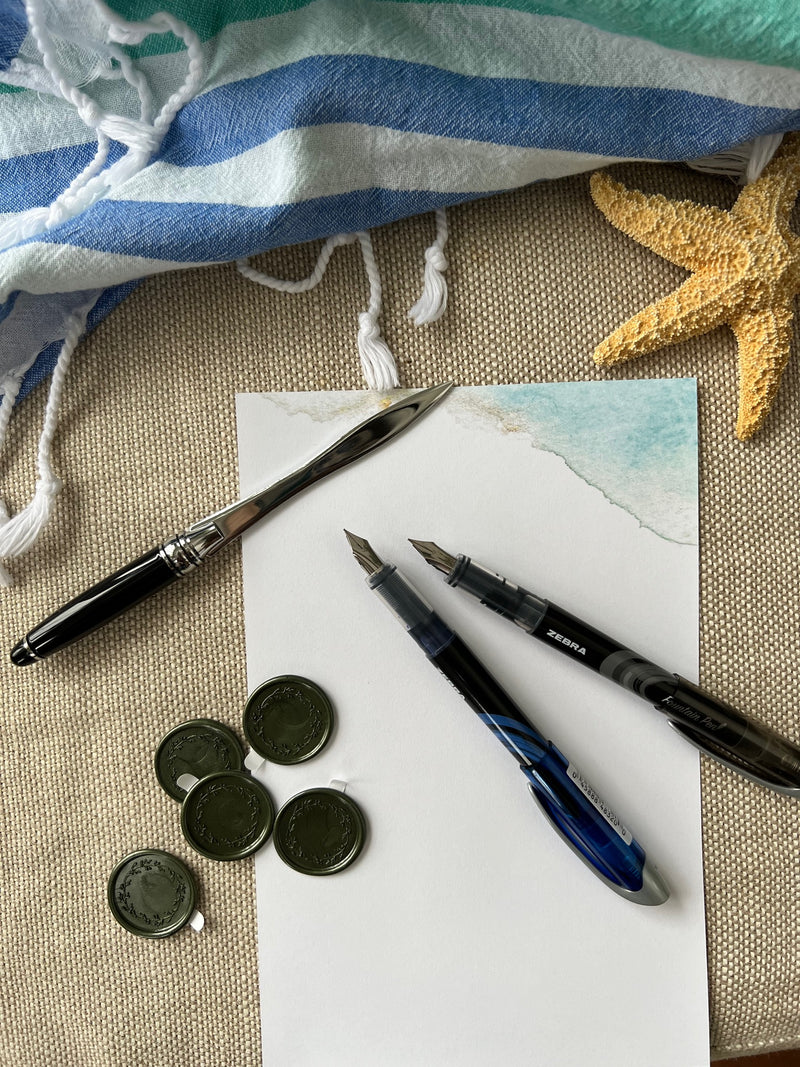 Letter opener, wax seals, 2 fountain pens and letter sheet surrounded by beach towel and starfish