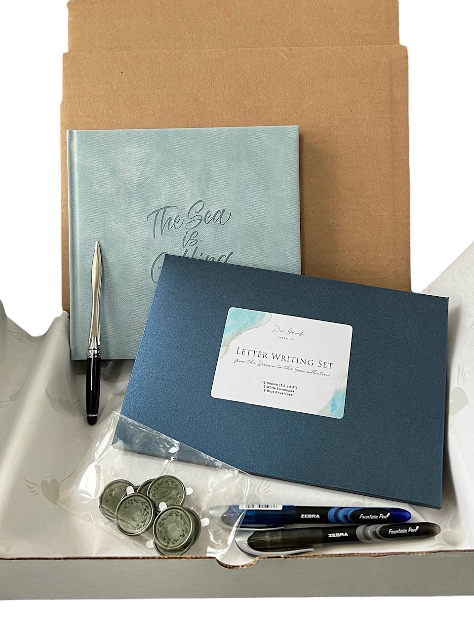 Subscription box items, such as a journal, letter writing set, pens, wax seals and a letter opener, displayed inside a box on a grey chair